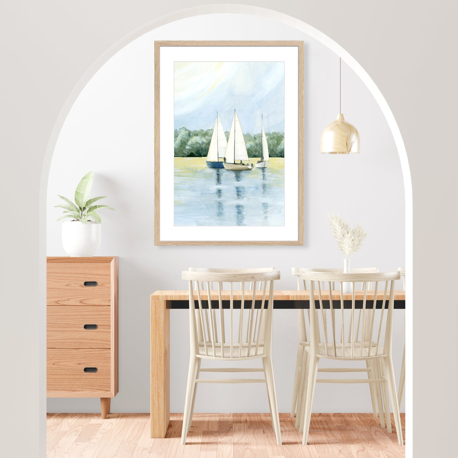Framed nautical artwork featuring sailboats on water, hanging in a minimalist dining room. The space includes a wooden table with white chairs, a light wood cabinet, and a plant under an arched doorway.