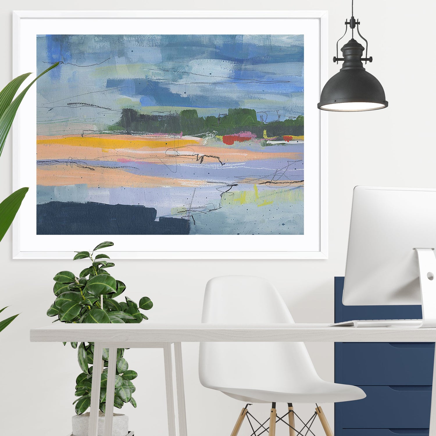 Framed abstract artwork with vibrant colours in blue, orange, and green tones hanging above a modern white desk in a minimalist home office. The space includes a white chair, a plant, and a black pendant light.