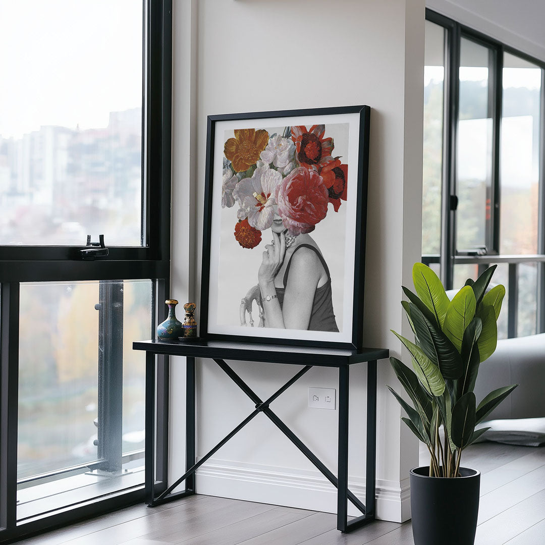 Framed floral portrait with colourful blooms over a black-and-white figure, displayed on a black console table in a bright modern room with large windows and a tall green plant.